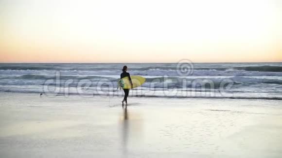 小女孩拿着冲浪板在海滩上女人带着海浪走进海洋美丽的夕阳风在吹视频的预览图