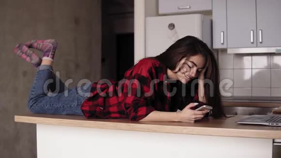 一个年轻女孩在她的厨房桌面上躺着发短信给某人时微笑和大笑的慢镜头视频的预览图