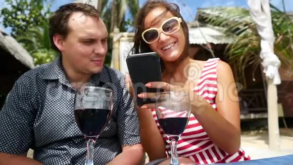 年轻快乐的夫妇喝葡萄酒使用智能手机在户外海滩咖啡馆休息慢动作1920x1080视频的预览图