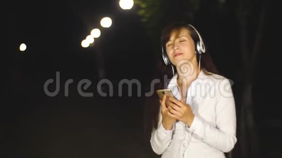 戴着耳机和智能手机的年轻女孩在一个城市公园里听音乐跳舞和微笑视频的预览图