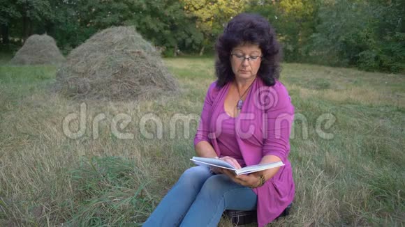 一位戴眼镜的老年妇女读了一本书坐在森林的空地上对着海斯塔克的背影视频的预览图