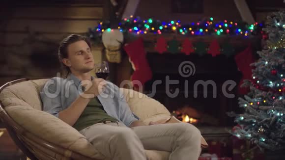 在圣诞夜一个长头发的男人坐在装饰的树旁端着一杯酒视频的预览图