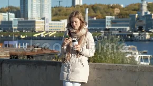 年轻的女人穿着白色夹克在河港喝着咖啡用电话微笑视频的预览图