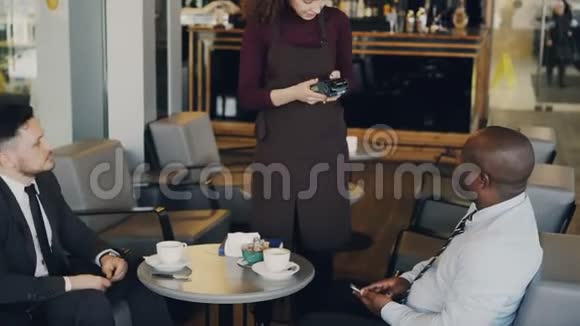 两个多种族的商人在正式服装支付在线账单使用智能手机在现代咖啡馆站着的年轻女服务员视频的预览图