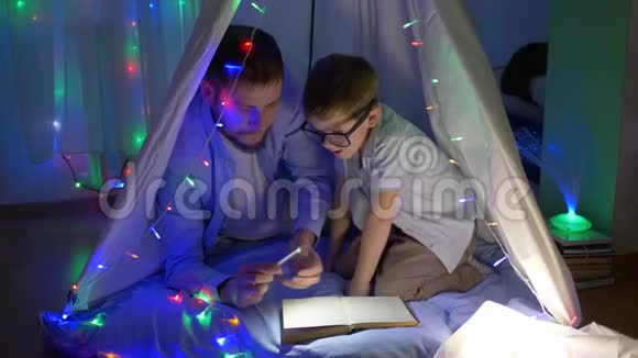 家庭教育爸爸拿着手电筒看书对着戴眼镜的孩子坐在帐篷里在黑暗中戴着花环视频的预览图