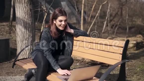 年轻漂亮的黑发女孩坐在公园的长凳上手提电脑里的人在挥手聊天笑着视频的预览图