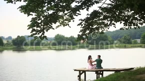 美丽的年轻夫妇坐在湖边的码头上浪漫的气氛背面视频的预览图