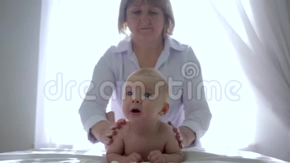 婴儿按摩穿白色实验室外套的女人在明亮的窗户背景下按摩一个小男孩的背部视频的预览图
