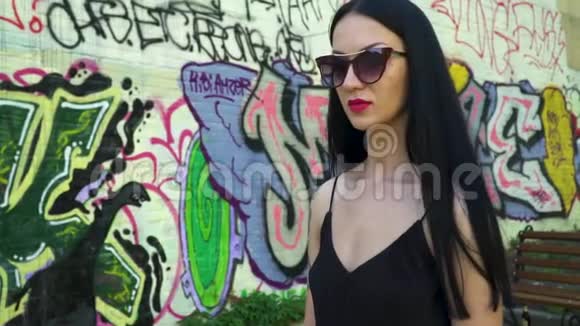 戴墨镜的黑发美女在涂鸦墙上慢悠悠地行走视频的预览图