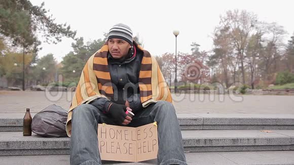 穿着破旧衣服的无家可归男子坐在城市公园里带着求救信号视频的预览图