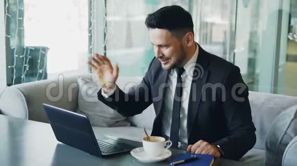 穿着正式服装的白人商人通过笔记本电脑网络摄像头聊天在一家高档咖啡馆里愉快地挥动着他的手视频的预览图