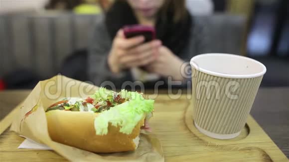 戴眼镜的学生女孩坐在咖啡馆里拿着智能手机一条多汁的开胃热狗就在前景中视频的预览图