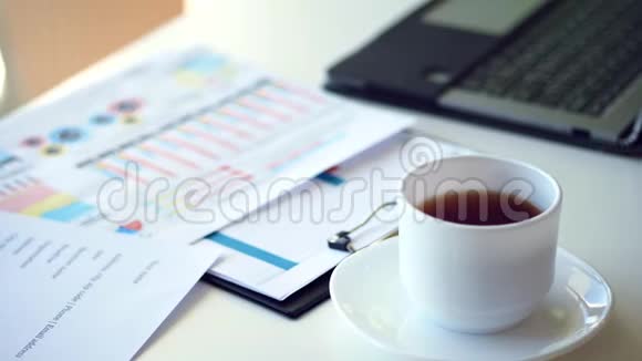 在一个白色的桌面上有一杯白色的茶旁边有一台笔记本电脑并展开商业图表视频的预览图