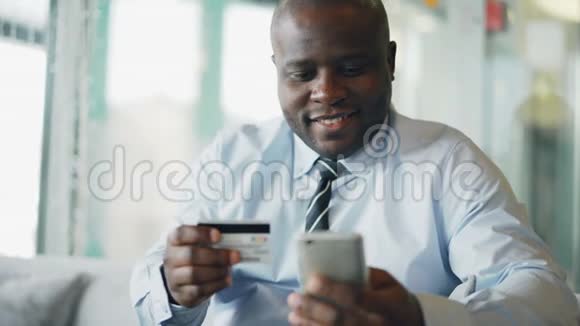 穿着正式服装的美国黑人商人在网上付账手里拿着信用卡和智能手机视频的预览图
