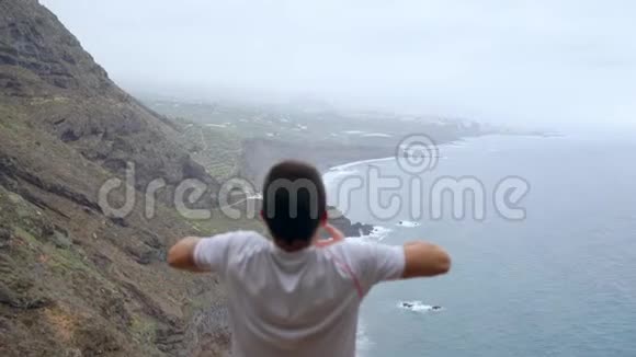 一个站在悬崖边上俯瞰大海的人举起双手在瑜伽的过程中吸入的空气视频的预览图