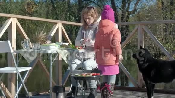 一个年轻的女人和一个穿暖和衣服的小女孩在烤架上煮蔬菜和肉一只狗在附近玩耍一只视频的预览图