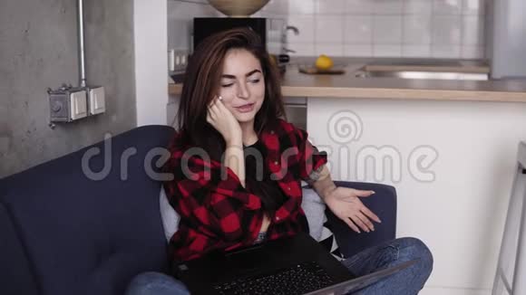 令人惊叹的美丽女孩与某人在电话中交谈而她的膝盖上拿着笔记本电脑坐在沙发上视频的预览图