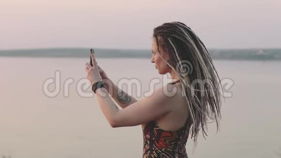迷人的嬉皮士女人带着可怕的家伙在海边日落时用智能手机拍照视频的预览图