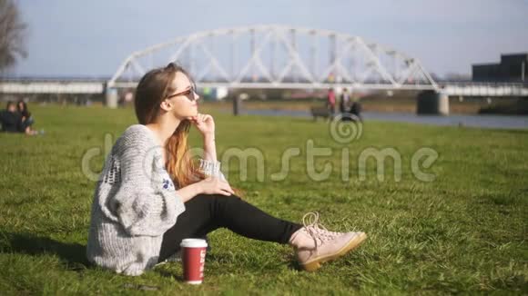 年轻的时髦女人坐在绿草上喝热咖啡早上享受新鲜空气享受外卖咖啡的女人视频的预览图