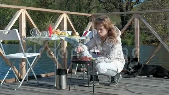 年轻女子穿着暖和的衣服在烤架上准备蔬菜和肉用电话狗在附近玩耍野餐视频的预览图