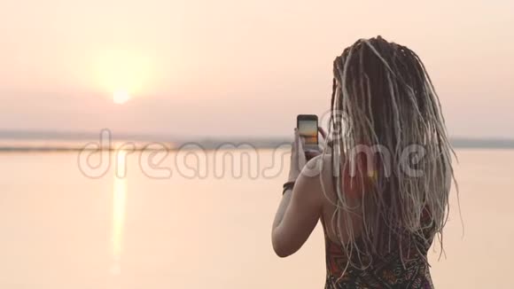 迷人的嬉皮士女人带着可怕的家伙在海边日落时用智能手机拍照视频的预览图