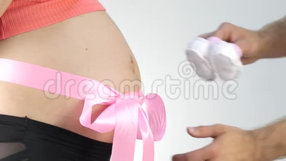男人手在玩婴儿鞋怀孕的腹部有粉红色的丝带4K视频的预览图