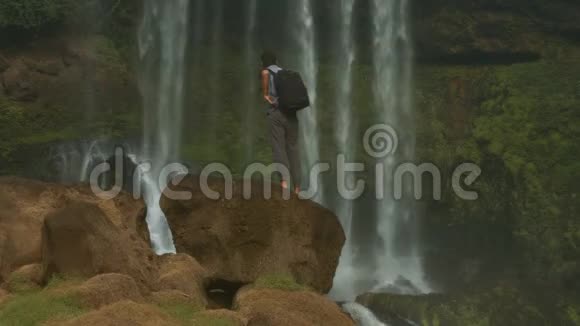 背包男站在岩石上喝着瀑布水视频的预览图