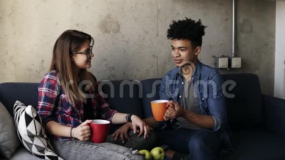 年轻的白种人女孩和英俊的穆拉特都穿着城市服装坐在沙发上喝茶说话视频的预览图