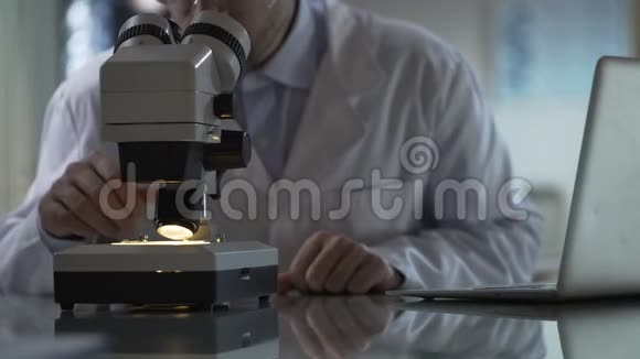 医学研究员在显微镜下观察样本在笔记本电脑上打字科学视频的预览图