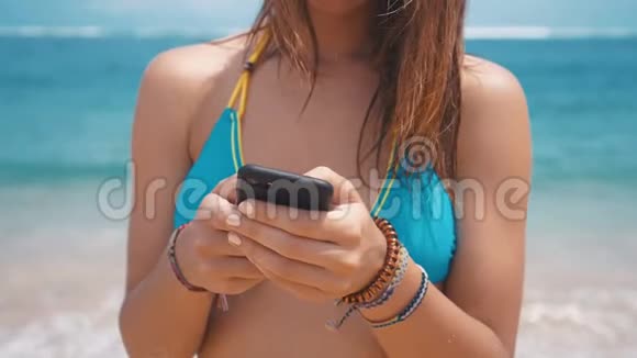 年轻女性使用智能手机在海滩的海洋背景微笑的女游客在巴厘岛度假时穿着泳衣视频的预览图