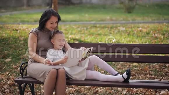 有雀斑的小女孩和妈妈坐在公园的长凳上一家人一起读一本书视频的预览图