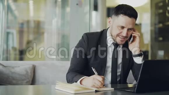 快乐的白人商人坐着微笑着和伴侣在智能手机上交谈并在记事本上写下信息视频的预览图