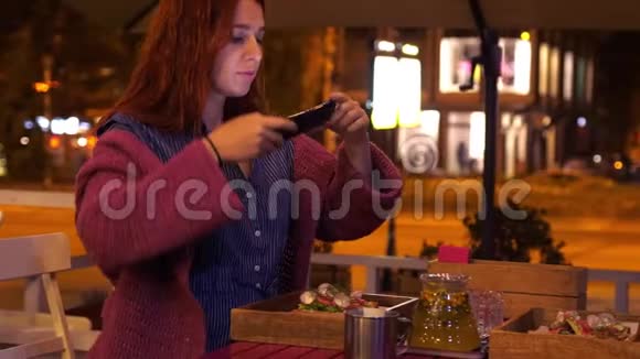 年轻漂亮的红头发女人在街边的咖啡馆里拍照路边有美丽的夜灯视频的预览图