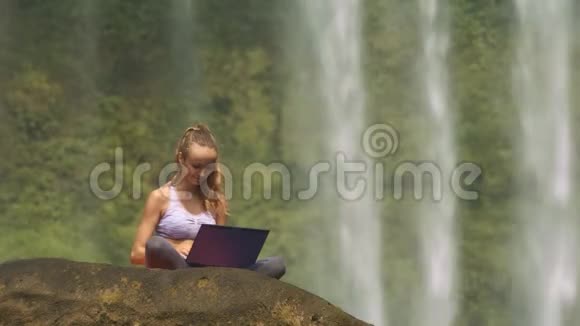 女孩把笔记本放在石头上在瀑布上打字视频的预览图