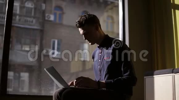 漂亮的年轻人坐在全景窗口附近在他的笔记本电脑上打字工作过程视频的预览图