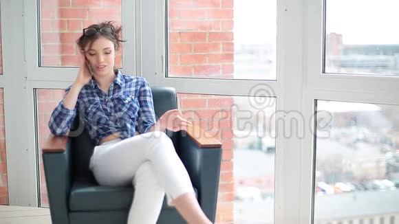 年轻女性使用智能手机打电话的肖像白人模特坐在靠近窗户的扶手椅上手里拿着视频的预览图