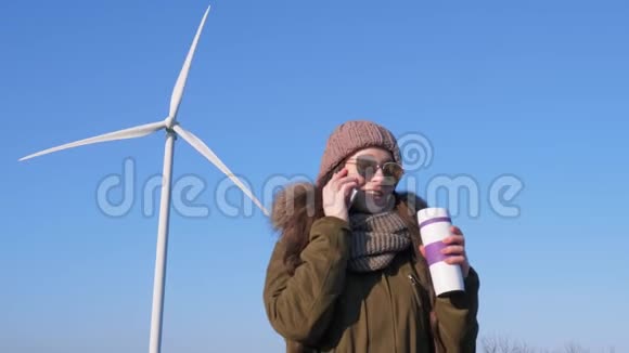 风力发电站戴眼镜的女性拿着热水瓶在对着天堂的风力涡轮机旁打电话视频的预览图