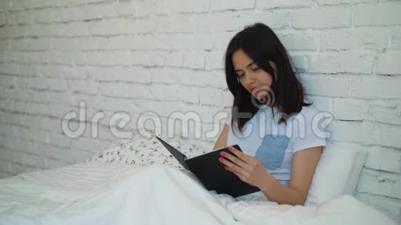 美丽的黑发女孩坐在床上拿着一本书看书一个年轻的女人打开一本有趣的书视频的预览图