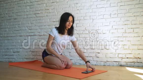 女人坐在健身房的瑜伽垫上看电话视频的预览图