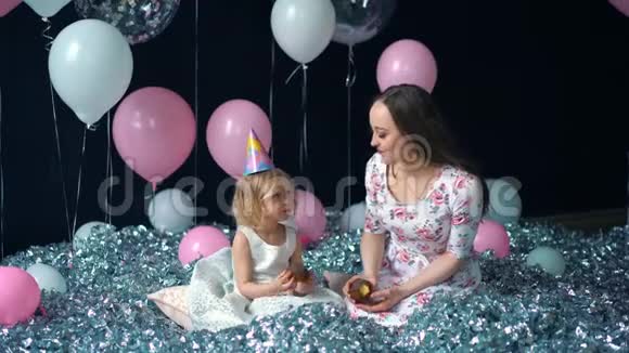 可爱的小女孩和她美丽的年轻妈妈吃纸杯蛋糕生日聚会节日聚会在气球的背景上视频的预览图