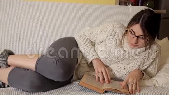 一个穿着白色毛衣的年轻女孩在一个冬天的晚上带着眼镜在家看一本有趣的书笑书晚上视频的预览图