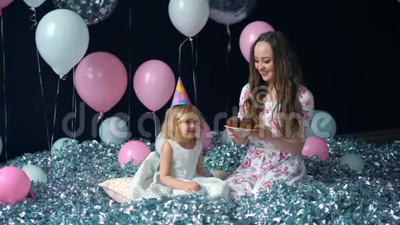 可爱的小女孩和她美丽的年轻妈妈吃纸杯蛋糕生日聚会节日聚会在气球的背景上视频的预览图