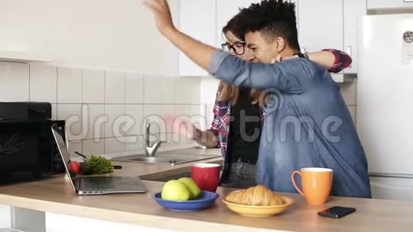 可爱的幸福夫妇在他们的笔记本电脑里向某人挥手视频聊天而在家里的厨房拥抱生活起居视频的预览图