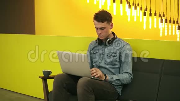 迷人的男性坐在沙发上在光线充足的室内空间喝咖啡在他的笔记本电脑上工作视频的预览图