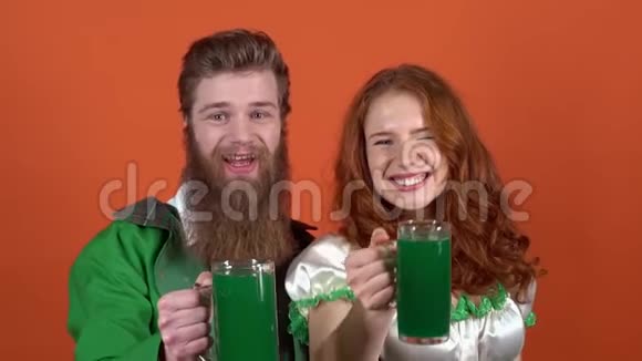 穿着圣诞服装的年轻一对夫妇庆祝圣帕特里克节他们被隔离在橙色的墙上提供绿色啤酒视频的预览图