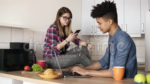 年轻的快乐夫妇坐在厨房里两人都使用一些设备如笔记本电脑智能手机而不是和他们说话视频的预览图