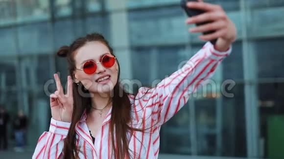 漂亮的年轻模特戴着红色太阳镜在她的智能手机上自拍一个漂亮的女孩站在购物旁边视频的预览图