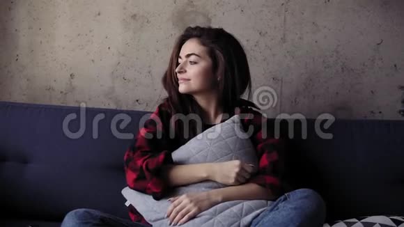 年轻漂亮的布鲁内特女孩坐在沙发上抱着枕头她试着打开电视但还记得视频的预览图