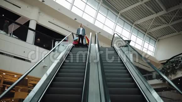 穿着牛仔裤夹克的年轻金发女人在购物中心的移动楼梯上倒下视频的预览图