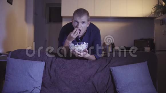 成人男子有兴趣观看电视和电影站在舒适的沙发附近手拿碗里吃美味的爆米花视频的预览图